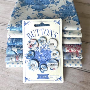May include: A set of eight fabric-covered buttons on a Tilda Special Edition 2026 card, featuring various blue and white floral, striped, and polka dot patterns. The buttons are arranged in a circular pattern, resting atop a stack of folded fabric.