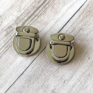 Pode incluir: Duas fivelas prateadas para bolsas. Cada fivela tem uma base arredondada, um centro em forma de U e uma peça superior com um pequeno detalhe redondo. As fivelas são feitas de metal brilhante e são projetadas para bolsas.
