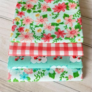 Peut inclure: Quatre carrés de tissu pliés avec différents motifs. Le tissu supérieur présente un motif floral bleu clair avec des fleurs roses et rouges. En dessous, un motif vichy rouge et blanc, suivi d'un tissu turquoise avec des fraises et des nuages, et un motif floral.