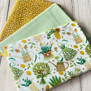 Könnte beinhalten: Drei Stoffmuster: ein senfgelber Stoff mit Punkten, ein hellgrüner Stoff mit Karomuster und ein weißer Stoff mit einem floralen und gartenbezogenen Druck. Der Druck umfasst Gießkannen, Topfpflanzen und Zitronen.