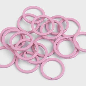 Puede incluir: Una colección de llaveros de color rosa claro. Los llaveros son circulares con una abertura con bisagras. Están hechos de un material sólido y tienen un acabado liso y mate. Los llaveros están dispersos sobre un fondo claro.