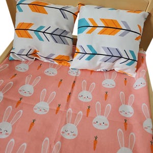 Peut inclure: Un lit de poupée en bois avec une literie à motif de lapin rose et blanc et deux oreillers à motifs de flèches orange, bleues et grises.