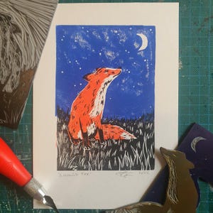 Peut inclure: Une linogravure d'un renard orange et blanc regardant le ciel nocturne. Le fond est bleu avec une lune en croissant et des étoiles. L'œuvre, intitulée "A Moonlight Fox" et datée de 2022, est imprimée sur papier.