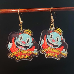 May include: A pair of dangle earrings featuring a cartoon character with a blue face, a yellow hat, and a red and black outfit. The earrings have the text "DON'T MAKE ME LAUGH..." in yellow. The earrings are suspended from gold-colored hooks.
