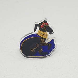May include: A black and gold acrylic charm featuring a stylized depiction of a creature with red eyes, a gold collar, and a blue cape. The creature is sitting on a blue and gold patterned background.