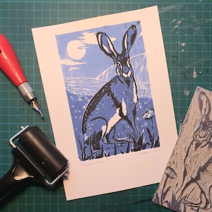 Op de afbeelding: Een blauwe en zwarte linosnede van een haas, getiteld "A Moorland Hare". De print is op een witte achtergrond en heeft een maan op de achtergrond.