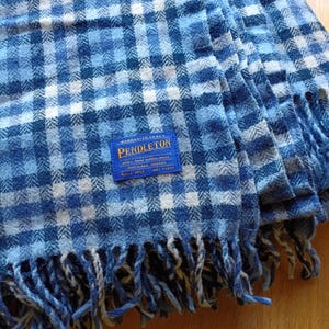 May include: A blue and white plaid wool blanket with fringe edges. A blue rectangular label is stitched onto the fabric, featuring the text Pendleton, 100% pure virgin wool, Portland, Oregon, and since 1863, with a small graphic of a sheep.
