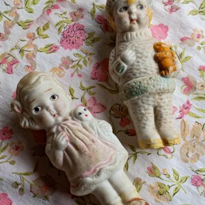May include: Two antique porcelain dolls. One doll wears a pink dress and has a small doll on its shoulder. The other doll wears a sweater and holds a small orange animal. The dolls are on a floral patterned fabric.