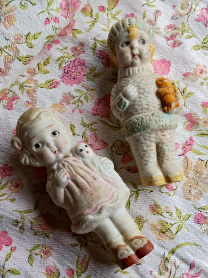 May include: Two antique porcelain dolls. One doll wears a pink dress and has a small doll on its shoulder. The other doll wears a sweater and holds a small orange animal. The dolls are on a floral patterned fabric.
