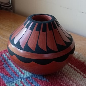 May include: A small, rounded terracotta vase featuring a smooth, polished finish. The exterior is decorated with a repeating black geometric pattern of vertical blades around the neck and a scalloped black band encircling the lower body.