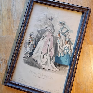 May include: Framed vintage print featuring three figures in period clothing. The print, titled "Journal des Demoiselles," is in a dark brown wooden frame. The artwork depicts women in long dresses and a young boy in a tunic.