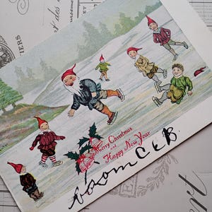 May include: A vintage postcard featuring a winter scene with several figures wearing red pointed hats and ice skates on a frozen pond. The text reads Merry Christmas and Happy New Year, with additional handwritten cursive script below the printed text.