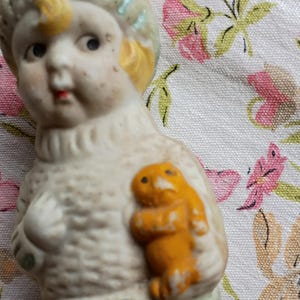 May include: A vintage porcelain figurine of a child with a yellow-haired head and a light blue hat. The figurine is holding a small orange figure. The figurine is on a floral patterned fabric.