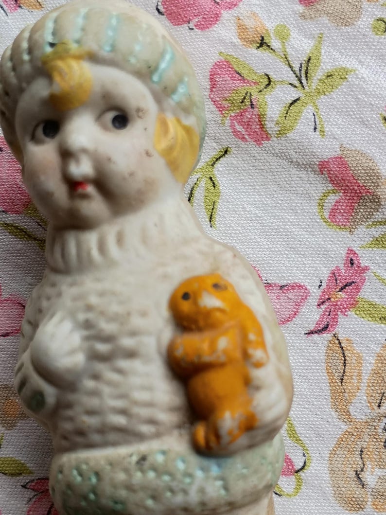 May include: A vintage porcelain figurine of a child with a yellow-haired head and a light blue hat. The figurine is holding a small orange figure. The figurine is on a floral patterned fabric.