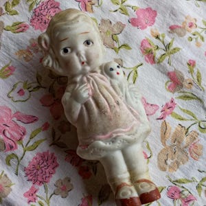 May include: A vintage porcelain doll in a pink dress, holding a small white dog figurine. The doll has blonde hair, blue eyes, and red accents on the lips and shoes. The doll is approximately 10 cm tall.