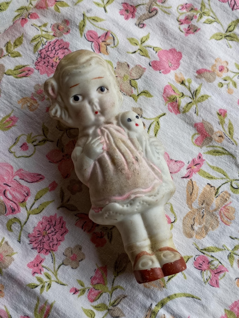 May include: A vintage porcelain doll in a pink dress, holding a small white dog figurine. The doll has blonde hair, blue eyes, and red accents on the lips and shoes. The doll is approximately 10 cm tall.