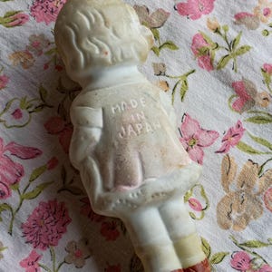 May include: A vintage porcelain doll figurine, viewed from the back, wearing a dress with a pink skirt and white top. The doll has blonde hair and is marked "MADE IN JAPAN" on the back. The doll's feet are painted red.