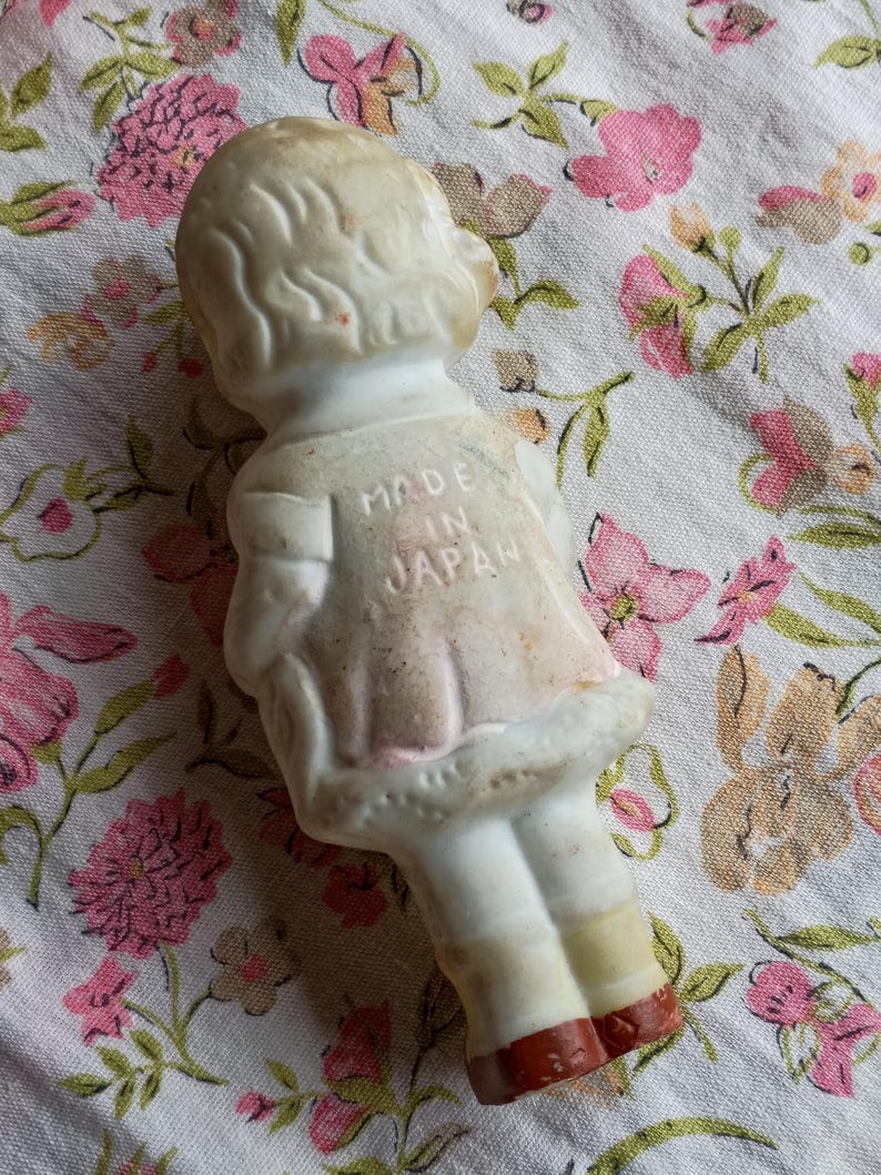 May include: A vintage porcelain doll figurine, viewed from the back, wearing a dress with a pink skirt and white top. The doll has blonde hair and is marked "MADE IN JAPAN" on the back. The doll's feet are painted red.