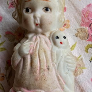 May include: A vintage bisque doll with blonde hair, blue eyes, and a red painted mouth. The doll is holding a smaller doll. The doll's dress has pink accents. The doll is a collectible item.