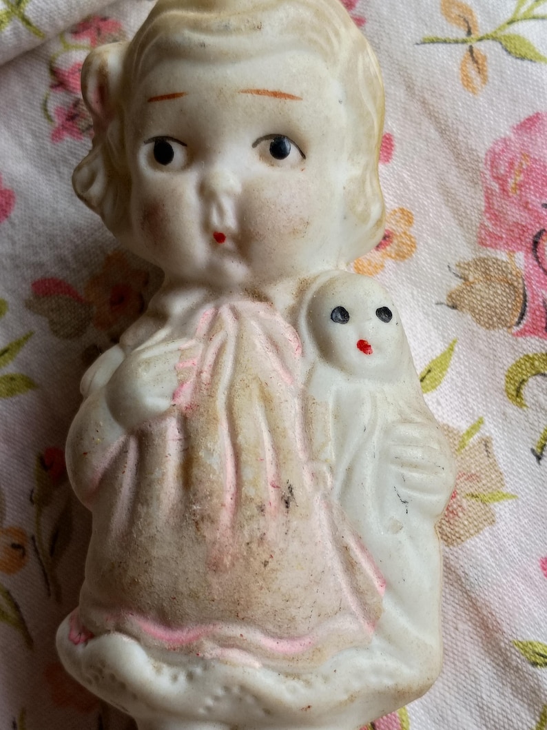 May include: A vintage bisque doll with blonde hair, blue eyes, and a red painted mouth. The doll is holding a smaller doll. The doll's dress has pink accents. The doll is a collectible item.
