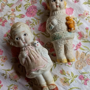 May include: Two antique porcelain dolls. One doll wears a pink dress and red shoes, holding a small white doll. The other doll wears a sweater and hat, holding a small orange animal. Both dolls have painted faces.