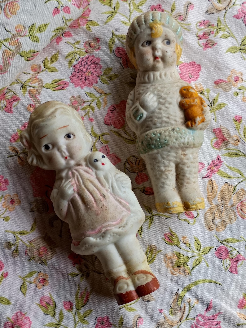 May include: Two antique porcelain dolls. One doll wears a pink dress and red shoes, holding a small white doll. The other doll wears a sweater and hat, holding a small orange animal. Both dolls have painted faces.