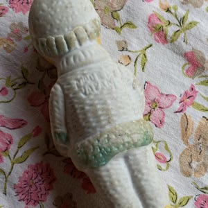 May include: A vintage ceramic doll figurine, viewed from the back, wearing a white outfit with green and yellow accents. The doll has a textured surface and the text "MADE IN JAPAN" is visible on the back.