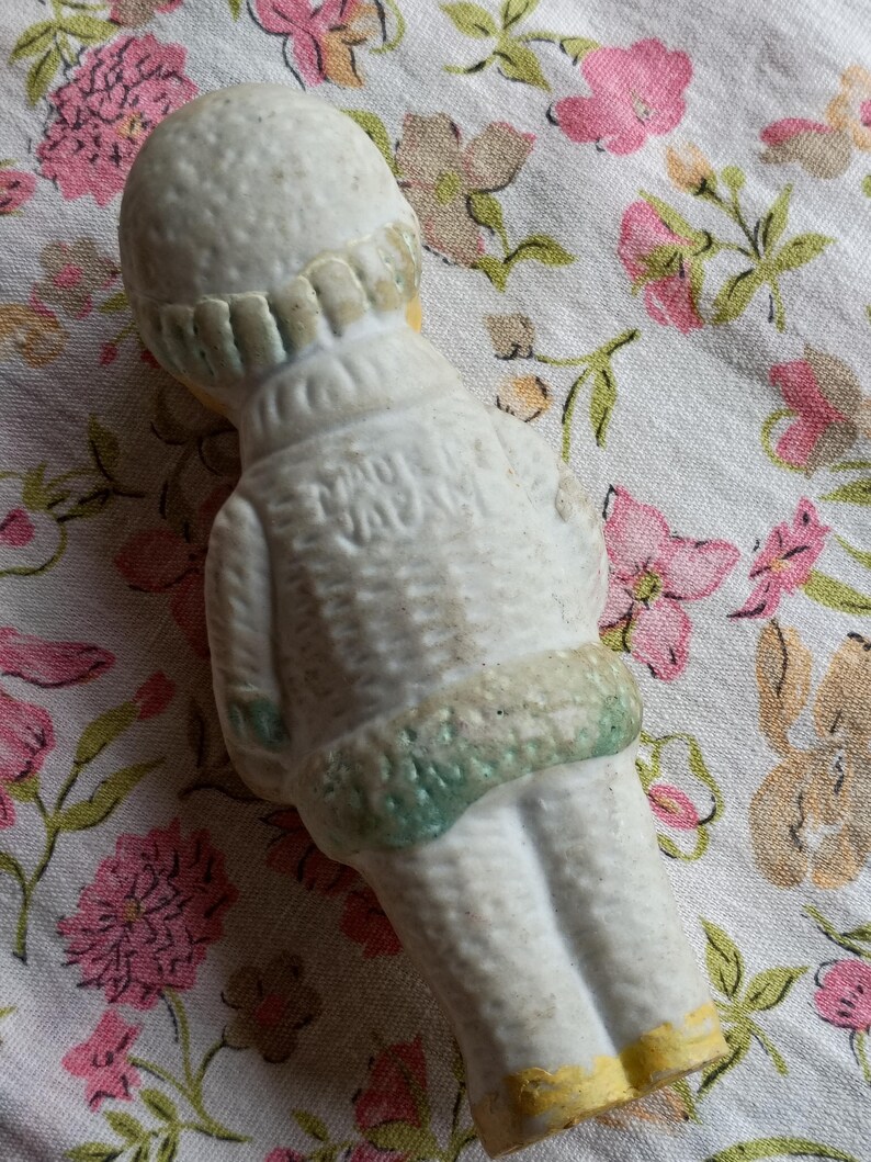 May include: A vintage ceramic doll figurine, viewed from the back, wearing a white outfit with green and yellow accents. The doll has a textured surface and the text "MADE IN JAPAN" is visible on the back.