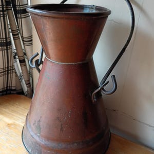 May include: A weathered copper milk can with a wide, flared top and a narrow neck. The vessel features a dark, oxidized patina with visible surface wear and a sturdy, arched metal handle attached to two side loops on the main body.