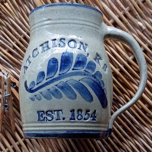 May include: A stoneware mug with a blue and white design. The mug features the text "ATCHISON KS" and "EST. 1854". The handle is curved, and the mug has a blue rim and base. The mug is sitting on a woven surface.