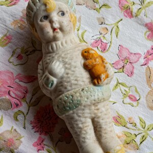 May include: A vintage bisque doll wearing a cream-coloured sweater and a light blue and yellow hat. The doll holds a small orange animal. The doll's face has blue eyes and a red mouth. The doll rests on floral patterned fabric.