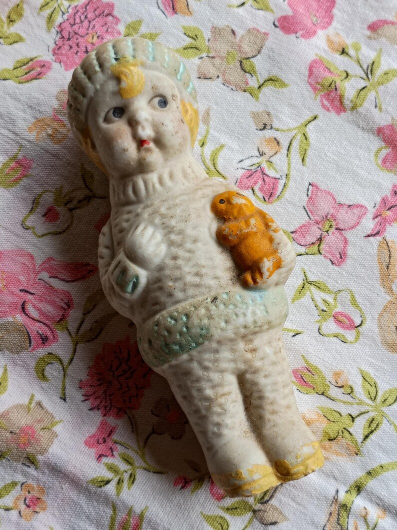 May include: A vintage bisque doll wearing a cream-coloured sweater and a light blue and yellow hat. The doll holds a small orange animal. The doll's face has blue eyes and a red mouth. The doll rests on floral patterned fabric.