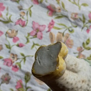 May include: Close-up of a ceramic figurine with a light grey surface and yellow detailing. The figurine is set against a floral patterned fabric with pink and brown flowers.