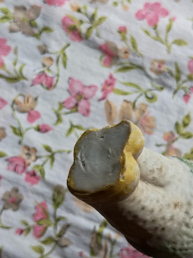 May include: Close-up of a ceramic figurine with a light grey surface and yellow detailing. The figurine is set against a floral patterned fabric with pink and brown flowers.