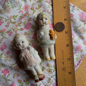 May include: Two vintage porcelain dolls with painted faces and detailed outfits. One doll holds a small bear, the other a small doll. They are on floral fabric, with a ruler and a penny nearby.
