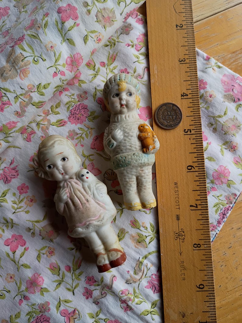 May include: Two vintage porcelain dolls with painted faces and detailed outfits. One doll holds a small bear, the other a small doll. They are on floral fabric, with a ruler and a penny nearby.