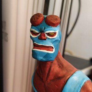 Puede incluir: Una figura de arcilla hecha a mano de un superhéroe con piel roja, una máscara azul y un mono azul. La figura tiene cuernos y una expresión seria.