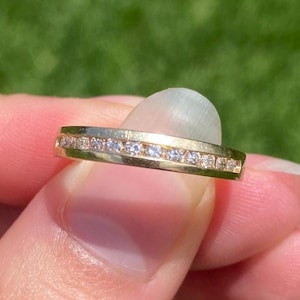 May include: A gold band ring featuring a channel setting with a row of small, clear, round-cut stones embedded along the center. The metal has a polished, reflective finish, and the stones are arranged in a continuous line across the front.