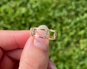 Puede incluir: Un anillo de banda dorada con un diseño abierto tipo jaula, formado por dos barras horizontales paralelas conectadas por tres segmentos verticales, cada uno engastado con pequeñas piedras claras y brillantes al frente.