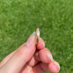 May include: A gold band ring featuring a channel set row of small, clear, sparkling stones along the center. The metal has a smooth, polished finish, and the ring is held upright to display the side profile and the embedded stones clearly.