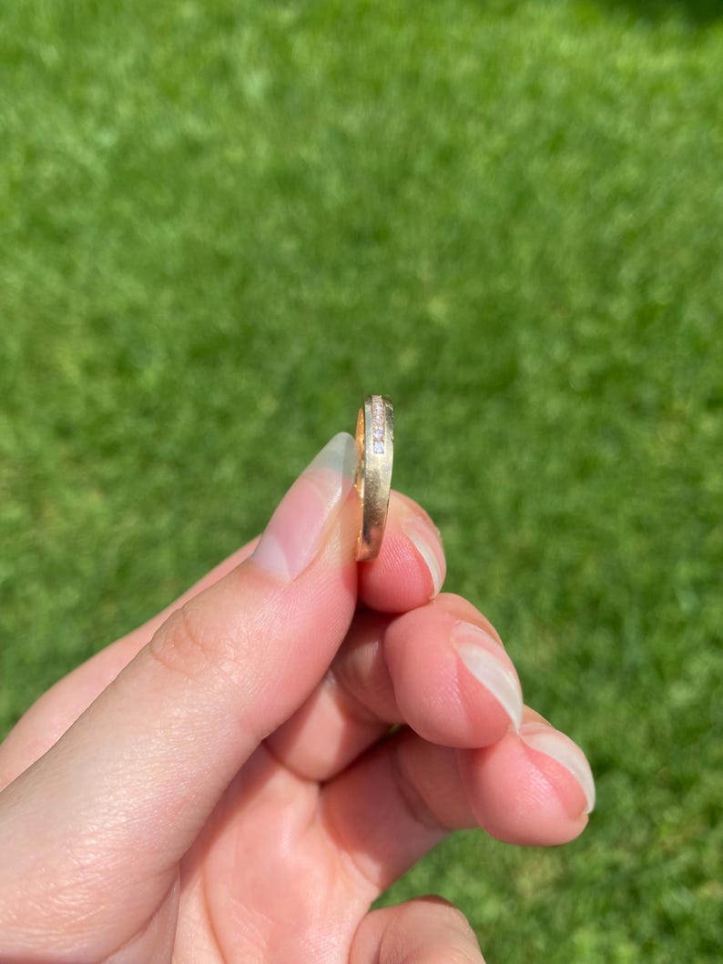 May include: A gold band ring featuring a channel set row of small, clear, sparkling stones along the center. The metal has a smooth, polished finish, and the ring is held upright to display the side profile and the embedded stones clearly.