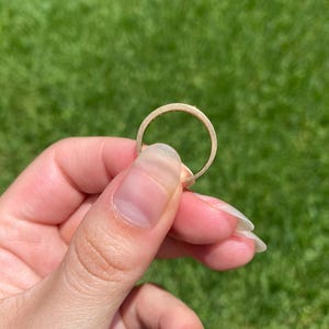 May include: A thin, circular gold ring with a matte, textured finish. The band has a consistent width and a smooth, rounded profile, showcasing a metallic yellow hue that reflects light across its surface in a simple, unadorned design.
