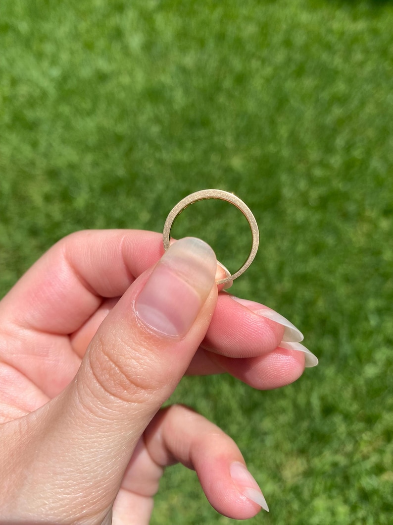 May include: A thin, circular gold ring with a matte, textured finish. The band has a consistent width and a smooth, rounded profile, showcasing a metallic yellow hue that reflects light across its surface in a simple, unadorned design.