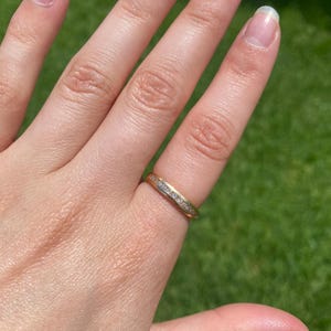May include: A gold band ring featuring a central channel set with a row of small, clear, round-cut stones. The ring has a polished metallic finish and a uniform width, showcasing a classic design with a continuous line of sparkling stones.