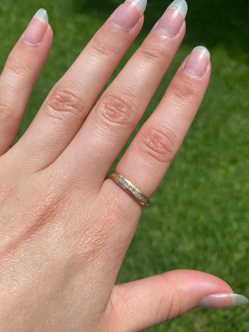 May include: A gold band ring featuring a central channel set with a row of small, clear, round-cut stones. The ring has a polished metallic finish and a uniform width, showcasing a classic design with a continuous line of sparkling stones.