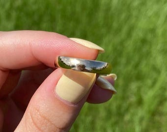 Puede incluir: Un anillo de banda de oro pulido con una superficie lisa y reflectante. El anillo presenta un perfil simple y redondeado con un diseño circular continuo, mostrando un acabado metálico brillante que capta la luz en toda su circunferencia exterior.