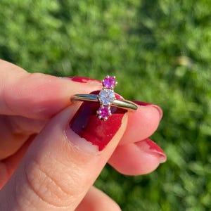 May include: A gold ring featuring a central clear gemstone flanked by two smaller pink gemstones. The ring is held up against a blurred green background, showcasing the ring's design and color contrast.