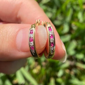 May include: A pair of gold hoop earrings with alternating red and clear gemstones. The earrings are held up against a blurred green background. The gemstones are set in a row along the front of the hoops.