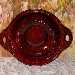 May include: A ruby red, oval-shaped glass serving dish with two handles. The dish has a textured rim and a detailed center design. The background features a floral pattern.