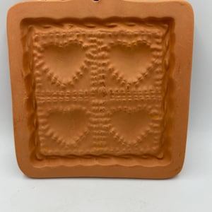 May include: A terracotta-coloured, square-shaped mould with a decorative heart pattern. The mould has a raised border and a small hole at the top for hanging. The heart design is repeated four times within the square.